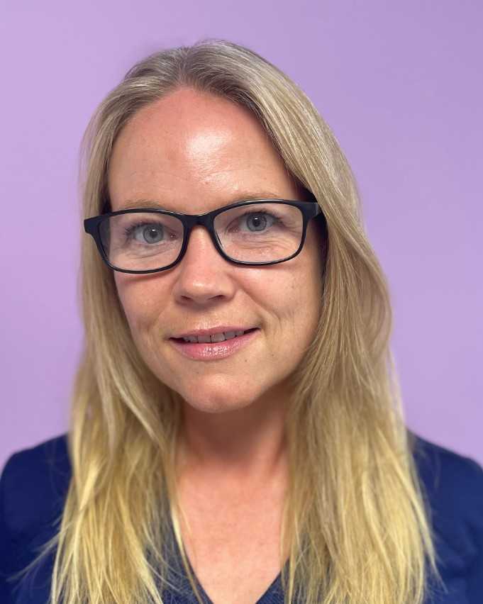 Portrett av en kvinne med langt blondt hår, svarte briller og et vennlig smil, iført en mørkeblå topp mot en lys lilla bakgrunn.