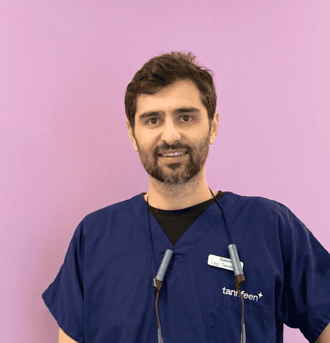 En mann med kort mørkt hår og skjegg står foran en rosa vegg, iført blå medisinske scrubs og stetoskop rundt halsen, og smiler mot kameraet.